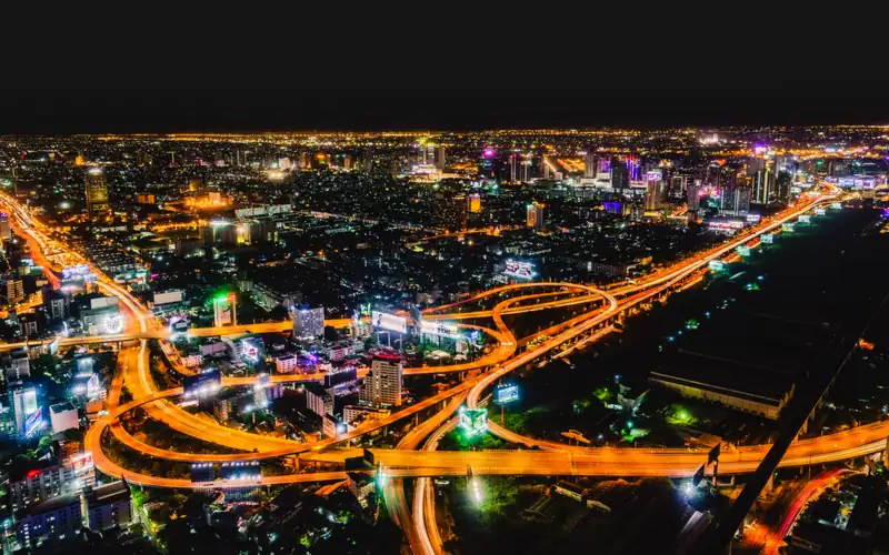 Bangkok at night from high above: glowing orange ribbons of an expressway interchange loop between lit buildings, and the city's lights stretch to the horizon under a dark sky